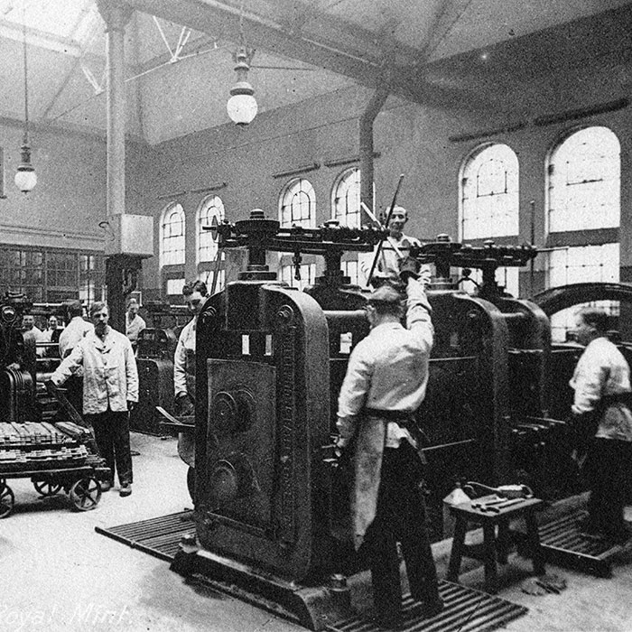 A black and white photograph of workers on the factory floor including two men working a coin press and one man wheeling a trolley full of coins.